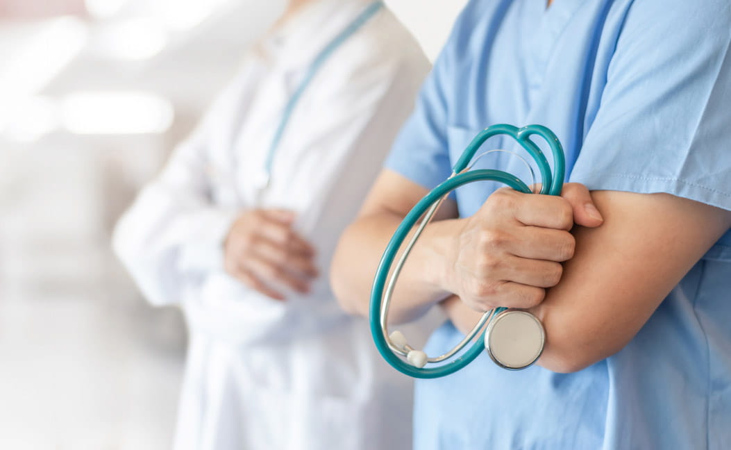 Stethoscope seen in the hand of a hospital professional wearing scrubs. A doctor in a white coat can be seen in the background.